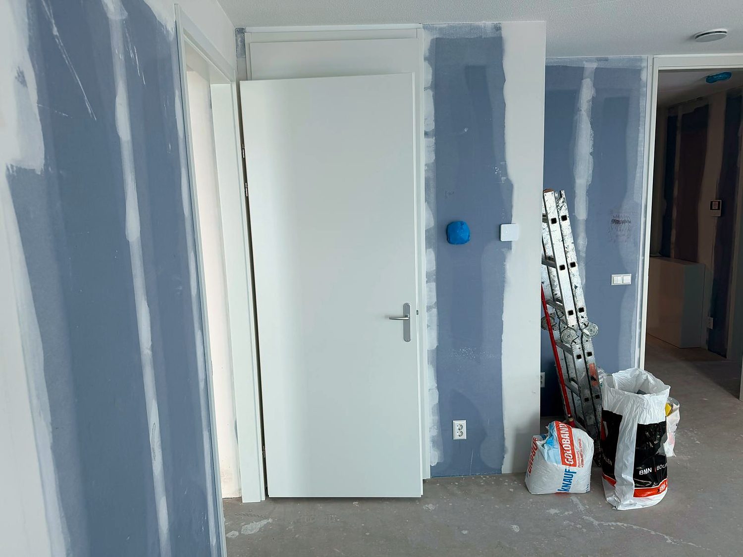 Nieuw geplaatste binnendeur in een gipswand tijdens de afbouw, met zakken stucmortel op de vloer