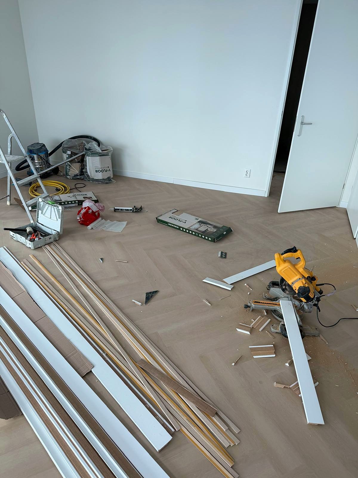 Visgraatvloer die gelegd wordt; de plinten, een verstekzaag en het gereedschap staan klaar in de ruimte