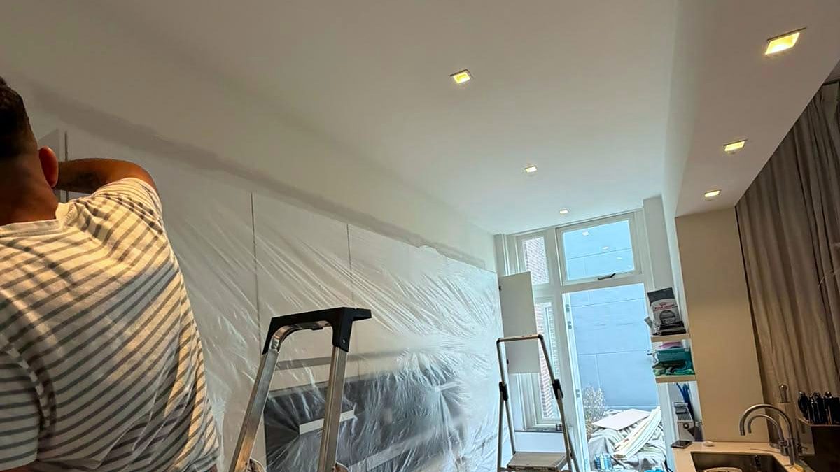 Een medewerker van MA LAYAN werkt op een trap aan de wandafwerking in een bewoonde woning; de keuken en de vloer zijn afgedekt met folie