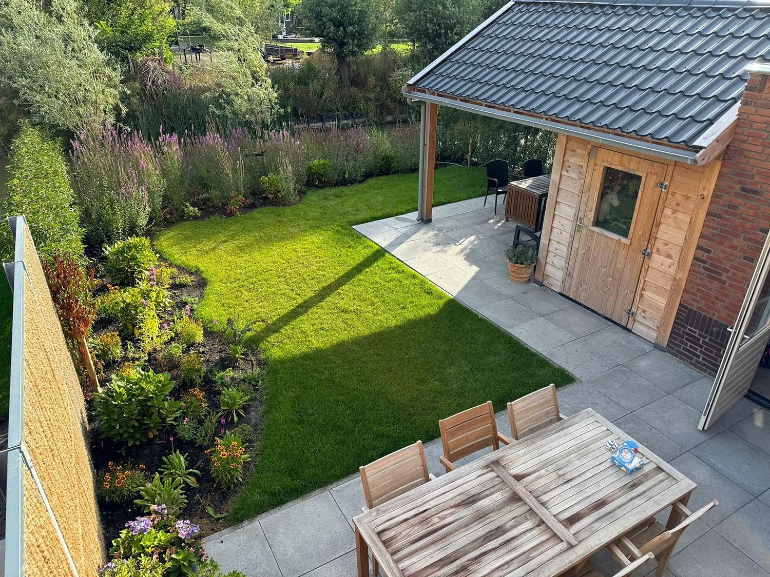 Opgeleverde tuin met een terras van grootformaat tegels, een strak gazon, beplanting langs het water en een houten berging onder een overkapping